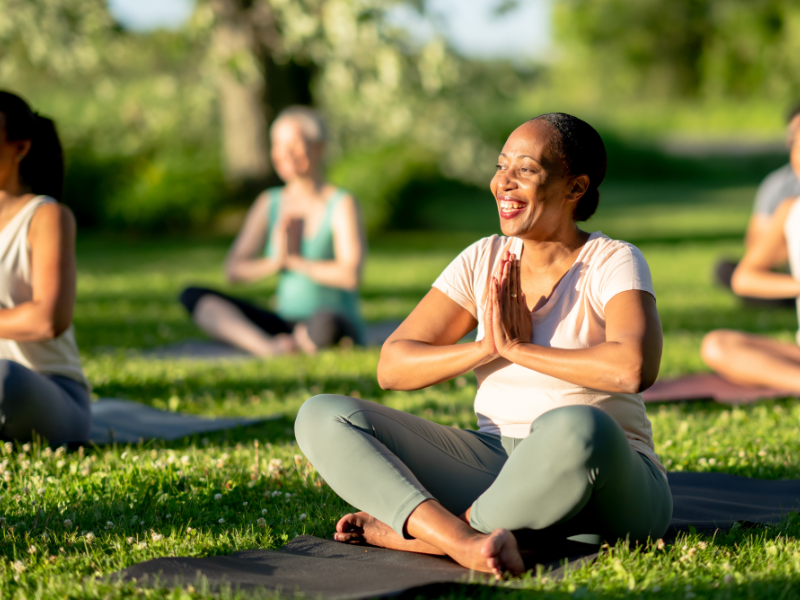 Gruppe von Menschen praktiziert Yoga im Freien auf einer grünen Wiese. Lächelnde Frau im Schneidersitz im Vordergrund.