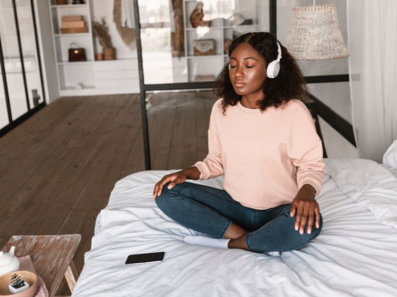 Eine junge Frau sitzt im Schneidersitz auf einem Bett und meditiert mit geschlossenen Augen. Sie trägt Kopfhörer und hört Musik. Das Schlafzimmer ist modern eingerichtet, mit einem hölzernen Nachttisch und einer dekorativen Hängelampe. bereit für die beste meditation