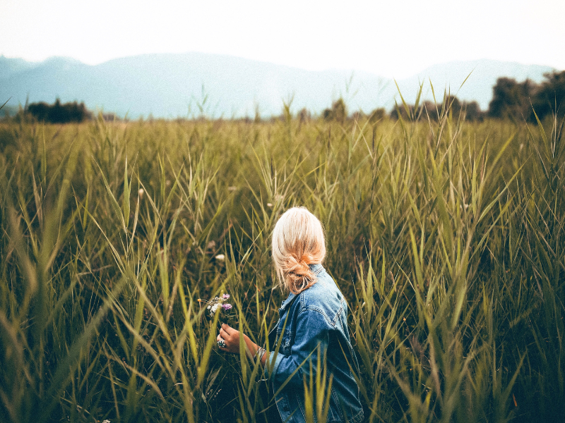 Eine Frau mit blonden Haaren, die in einem lockeren Knoten gebunden sind, sitzt in einem hohen Feldgras und hält einen kleinen Blumenstrauß in der Hand. Im Hintergrund sind Berge in der Ferne zu sehen.