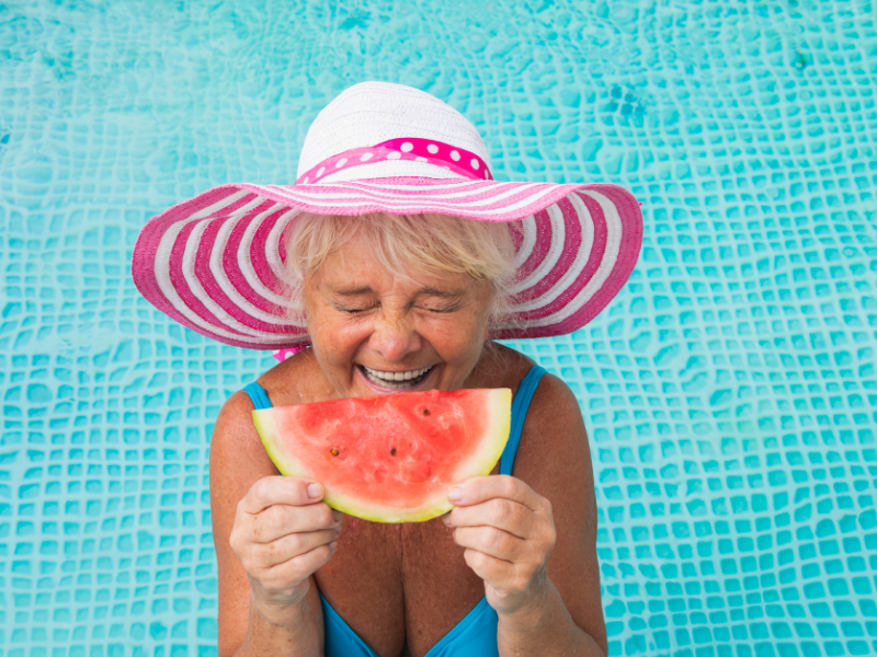 Eine fröhliche ältere Frau mit einem gestreiften Sonnenhut isst lachend ein Stück Wassermelone am Pool. Der Hintergrund zeigt das klare, blaue Wasser des Pools.