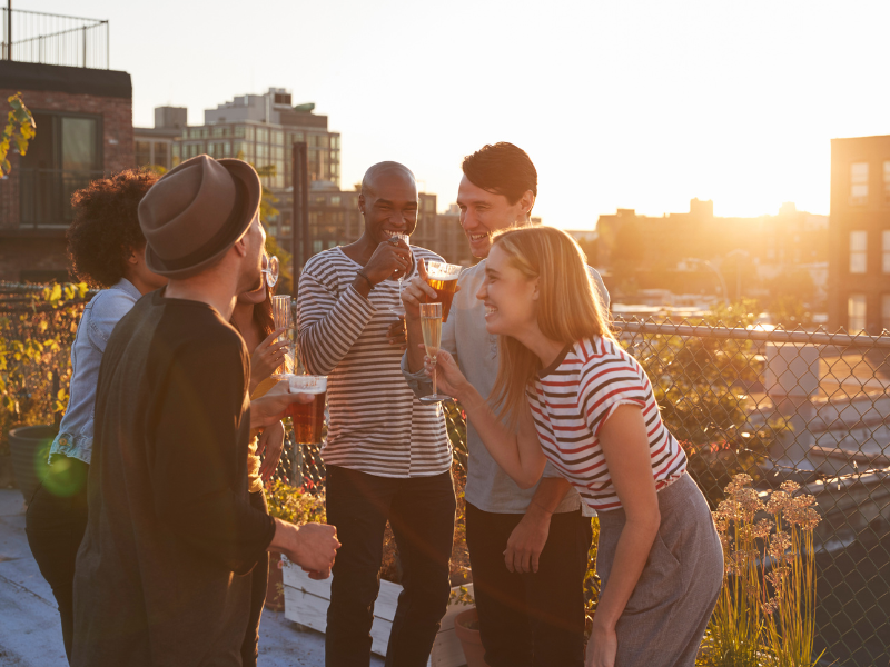 Eine Gruppe von Freunden genießt eine Rooftop-Party bei Sonnenuntergang. Sie lachen, trinken und haben eine gute Zeit auf der Dachterrasse inmitten einer urbanen Umgebung