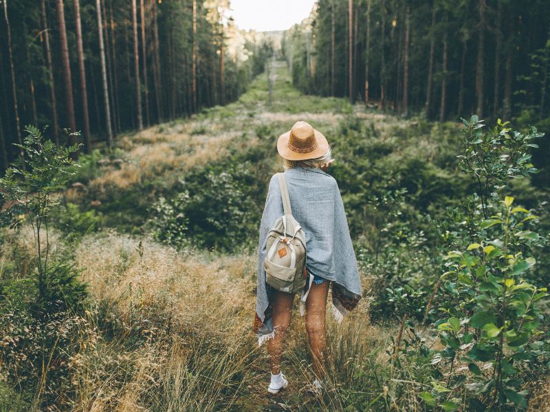 Selbstfürsorge und Selbstliebe durch Waldbaden. Frau mit Hut und Rucksack steht am Rand eines Waldes und blickt in die Ferne, symbolisiert die Erkundung und Entschleunigung im Einklang mit der Natur durch Slow Living.