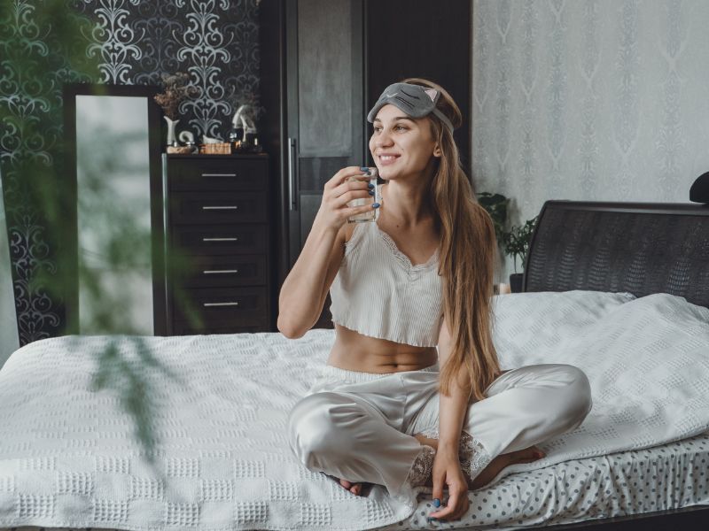 Selbstfürsorge Morgenroutine. Eine Frau sitzt auf ihrem Bett, trägt Schlafkleidung und hält eine Tasse, während sie entspannt lächelt.