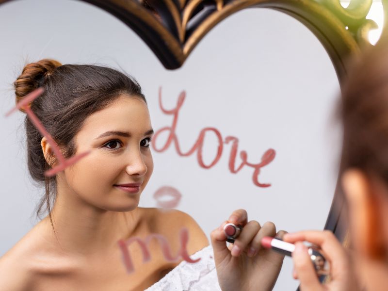 Selbstliebe üben. Eine Frau schreibt die Botschaft "I love me" mit Lippenstift auf einen Herz-förmigen Spiegel.