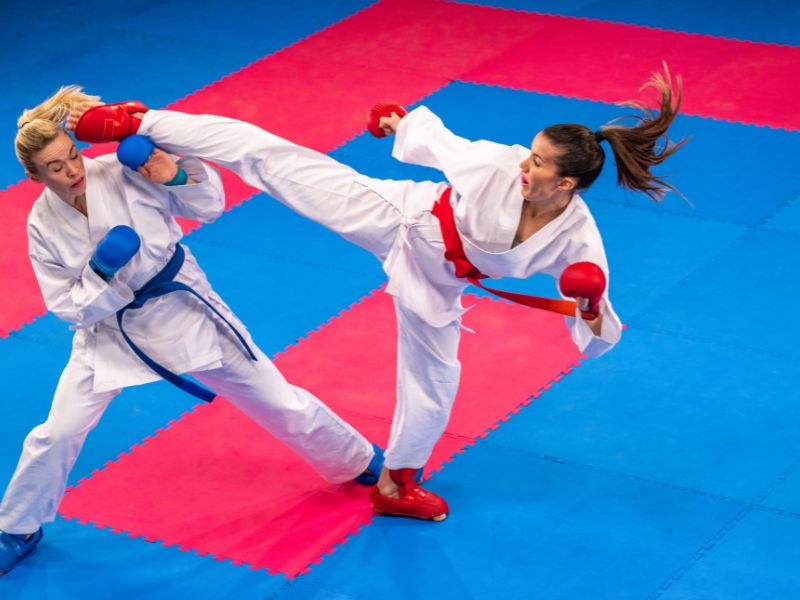 Zwei weibliche Karatekas im intensiven Kumite-Kampf, eine führt einen präzisen Tritt aus, während die andere eine Abwehrhaltung einnimmt. Perfekte Darstellung von Technik und Dynamik im Karate Kumite.