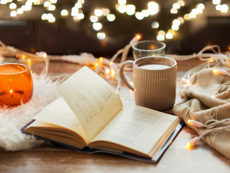 Offenes Buch mit Kerze und Tasse Kakao auf Holztisch, Lichterkette im Hintergrund – perfekte Hygge Idee für den Herbst